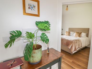 A table with a green plant in a rusty pot and a visitor book next to an open door showing a bedroom with a bed and pillows at Monks Rest in Monknash