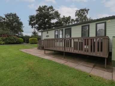 A garden with a lawn and bushes in front of a mobile home with a wooden deck at Dove in Shobdon