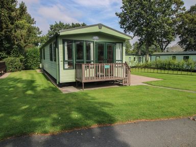 A small green cabin with a wooden porch and steps surrounded by grass and trees at Dove in Shobdon