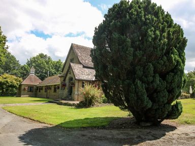A stone building with a steep roof and hanging flower baskets near a large bush on a lawn at Dove in Shobdon