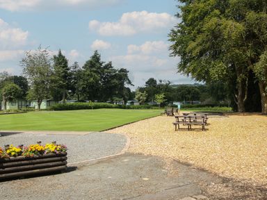 An outdoor area with a green lawn and wooden benches surrounded by trees and small buildings at Dove in Shobdon