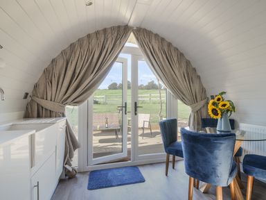 A dining area with blue chairs and sunflowers on a glass table near large windows with curtains at Cedar in Wootton Fitzpaine near Charmouth