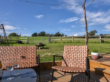 Two woven chairs on a wooden deck overlooking a grassy field with a wire fence and string lights above at Cedar in Wootton Fitzpaine near Charmouth