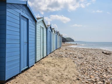 A row of blue beach huts on a rocky and sandy shore by the sea with cliffs in the background