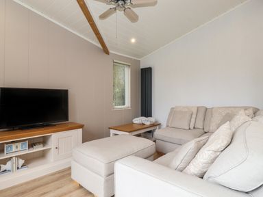 A living room with a television on a wooden cabinet a beige sofa and ottoman a wooden coffee table and a ceiling fan at Serendipity R11 Fallbarrow Park near Bowness-On-Windermere