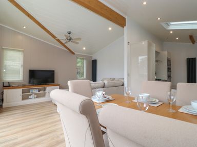 A dining area with beige chairs and a wooden table set with dishes and glasses a living room with a sofa a ceiling fan and a TV on a stand at Serendipity R11 Fallbarrow Park near Bowness-On-Windermere