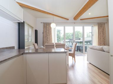 A kitchen area with a dining table and beige chairs next to a living room with a sofa and glass doors at Serendipity in R11 Fallbarrow Park near Bowness-On-Windermere