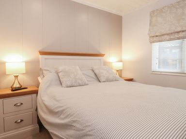 A bedroom with a bed flanked by two bedside tables with lamps and a window with blinds and a patterned curtain at Serendipity R11 Fallbarrow Park near Bowness-On-Windermere