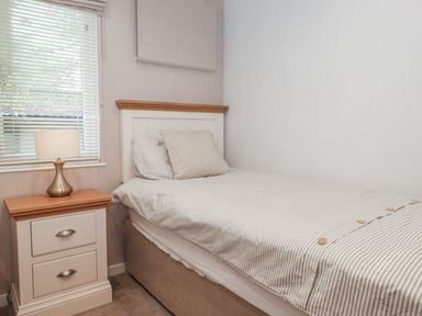 A single bed with striped bedding and a pillow beside a window with blinds and a nightstand with a lamp at Serendipity R11 Fallbarrow Park near Bowness-On-Windermere