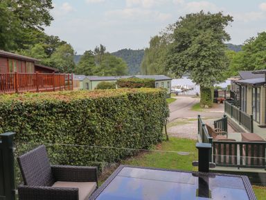 An outdoor seating area with a glass table and woven chair overlooking a hedge a pathway a tree and mobile homes near a lake at Serendipity in R11 Fallbarrow Park near Bowness-On-Windermere