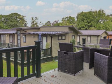 A balcony with dark wicker chairs and a glass table overlooking beige modern cabins and trees at Serendipity R11 Fallbarrow Park near Bowness-On-Windermere