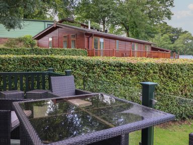 An outdoor seating area with a glass table and wicker chairs next to a hedge and wooden cabin at Serendipity R11 Fallbarrow Park near Bowness-On-Windermere