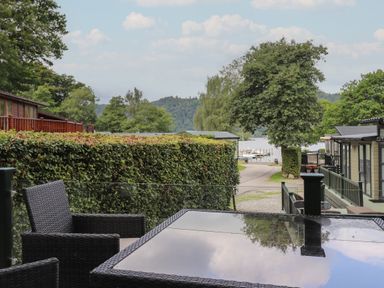 An outdoor patio with wicker chairs and a glass table overlooking a hedge garden and a lakeside area with boats at Serendipity in R11 Fallbarrow Park near Bowness-On-Windermere