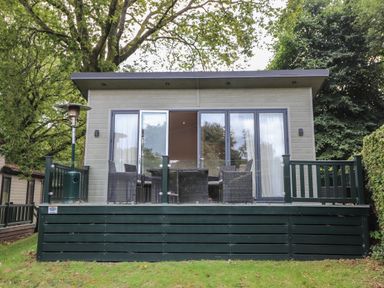 A small cabin with a deck featuring outdoor furniture and a heater surrounded by trees at Serendipity R11 Fallbarrow Park near Bowness-On-Windermere