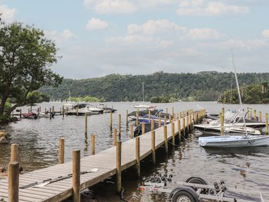 A wooden dock with several boats and a small trailer on a lake with forested hills in the background at Serendipity R11 Fallbarrow Park near Bowness-On-Windermere