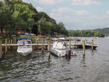 A dock with multiple boats moored on a lake with trees and houses in the background at Serendipity R11 Fallbarrow Park near Bowness-On-Windermere