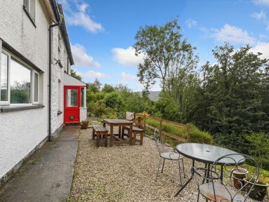 An outdoor patio area with wooden picnic table and benches a round glass table with metal chairs and a red door on a white building at Delfryn in Goginan near Aberystwyth