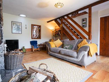 A living room with a gray sofa yellow cushions a wooden staircase and baskets with firewood at Delfryn Goginan in Goginan near Aberystwyth