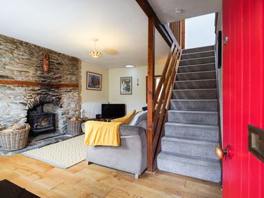 A living room with a stone fireplace and wood stove a gray sofa with a yellow blanket a television and carpeted stairs at Delfryn  Goginan in Goginan near Aberystwyth