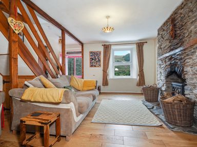 A living room with a sofa yellow cushions a wooden side table a stone fireplace and windows with curtains at Delfryn Goginan in Goginan near Aberystwyth