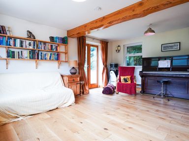A living room with a bookshelf a covered sofa a wooden cabinet a red armchair a piano and wooden flooring at Delfryn Goginan in Goginan near Aberystwyth