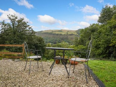A patio area with a round metal table and two metal chairs overlooking trees and hills at Delfryn Goginan in Goginan near Aberystwyth