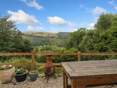 A garden patio with potted plants a chiminea and a wooden table overlooking hills and trees at Delfryn Goginan in Goginan near Aberystwyth