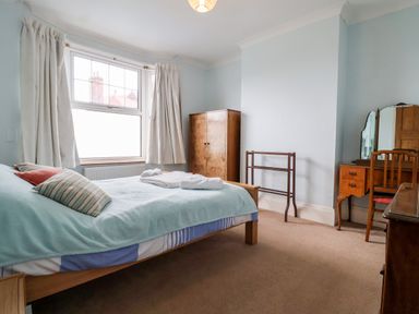 A bedroom with a bed with pillows and towels near a window with curtains a wardrobe a dressing table with mirror and chair at The Pink House in Felixstowe Suffolk