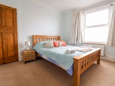 A bedroom with a wooden bed, bedside table, lamp, window with curtains and towels on the bed at The Pink House in Felixstowe Suffolk