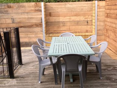 An outdoor patio with a light blue wooden table and six gray plastic chairs surrounded by wooden fencing