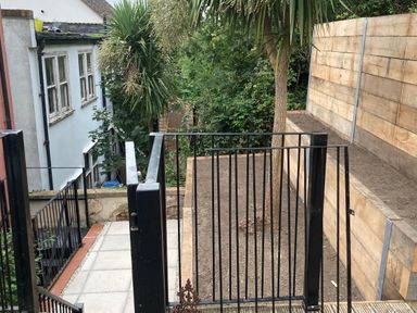 A garden area with a tree fenced by a black metal gate and wooden retaining walls next to a paved path at The Pink House in Felixstowe Suffolk