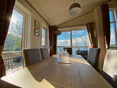 A dining room with a wooden table and gray chairs overlooking a lake through large windows at Pine Lake View in Coniston View 3 Carnforth