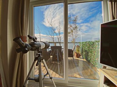 A telescope in front of glass doors leading to a deck with chairs and a table overlooking a lake at Pine Lake View in Coniston View 3 Carnforth