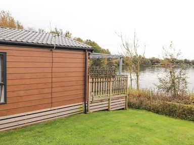 A brown wooden house next to a wooden fence with a lake and trees in the background at Pine Lake View in Coniston View 3 Carnforth