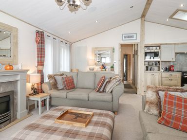 A living room with beige sofas and plaid cushions near a fireplace and an open kitchen at Pine Lake View in Coniston View 3 Carnforth