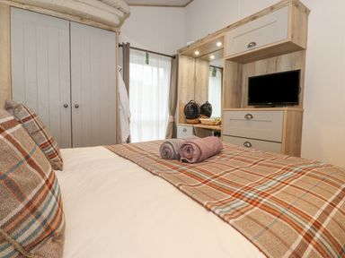 A bedroom with a bed covered in plaid blankets and pillows two rolled towels on the bed a wardrobe a window with curtains a dresser with a mirror and a television at Pine Lake View in Coniston View 3 Carnforth