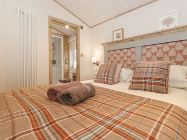 A bedroom with a bed with plaid bedding and pillows two rolled towels on the bed a wall lamp and an ensuite bathroom at Pine Lake View Coniston View 3 Carnforth