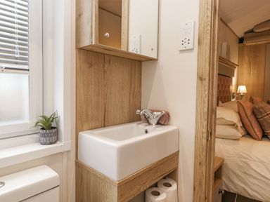 A small bathroom sink with a mirror cabinet above and toilet paper below next to a window with blinds and a bedroom visible through a wooden doorway at Pine Lake View Coniston View 3 Carnforth