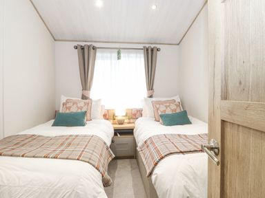 A bedroom with two single beds separated by a nightstand with a lamp and curtains on the window at Pine Lake View in Coniston View 3 Carnforth