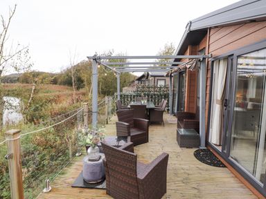 An outdoor wooden deck with wicker chairs a table with a glass top and a railing overlooking vegetation at Pine Lake View Coniston View 3 Carnforth