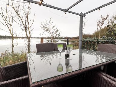 An outdoor dining area with a glass table two green-tinted wine glasses and a wine bottle overlooking a lake at Pine Lake View in Coniston View 3 Carnforth