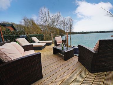 An outdoor deck with wicker chairs and loungers overlooking a lake at Pine Lake View in Coniston View 3 Carnforth
