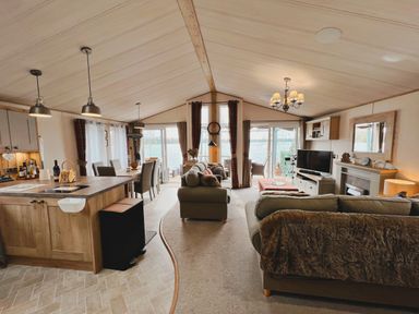 An open living area with a kitchen island dining table sofas and a television by large windows at Pine Lake View in Coniston View 3 Carnforth