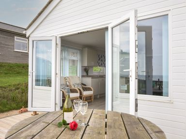 An open doorway leading to a room with armchairs and a kitchen area with flowers on the counter a wooden table with a bottle of wine two glasses and a red rose outside at Sealark in Eype