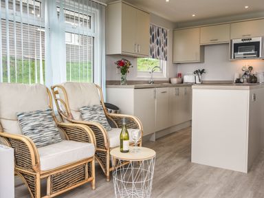 A kitchen with cabinets a microwave a sink and two wicker armchairs with cushions and a small table with a bottle and glasses at Sealark in Eype