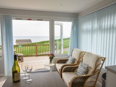 Two wicker chairs with cushions and pillows next to a small table with flowers in a bright room with large windows and a view of grass and sea at Sealark in Eype