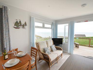 A dining table set for two next to a wicker loveseat with cushions by windows overlooking a deck and sea at Sealark in Eype