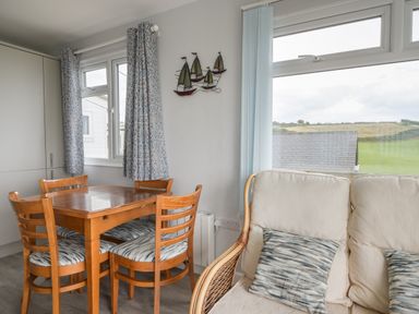 A dining area with a wooden table and four chairs next to a white cushioned sofa with patterned pillows and windows showing an outdoor view at Sealark in Eype
