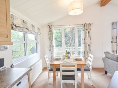 A dining area with a wooden table set for four near windows with floral curtains and a grey sofa at The Hideaway in Eype