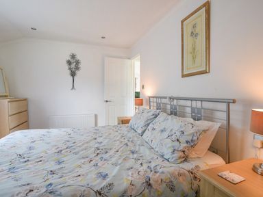 A bedroom with a bed featuring floral bedding a wooden dresser a bedside table with a lamp and floral artwork on the wall at The Hideaway in Eype